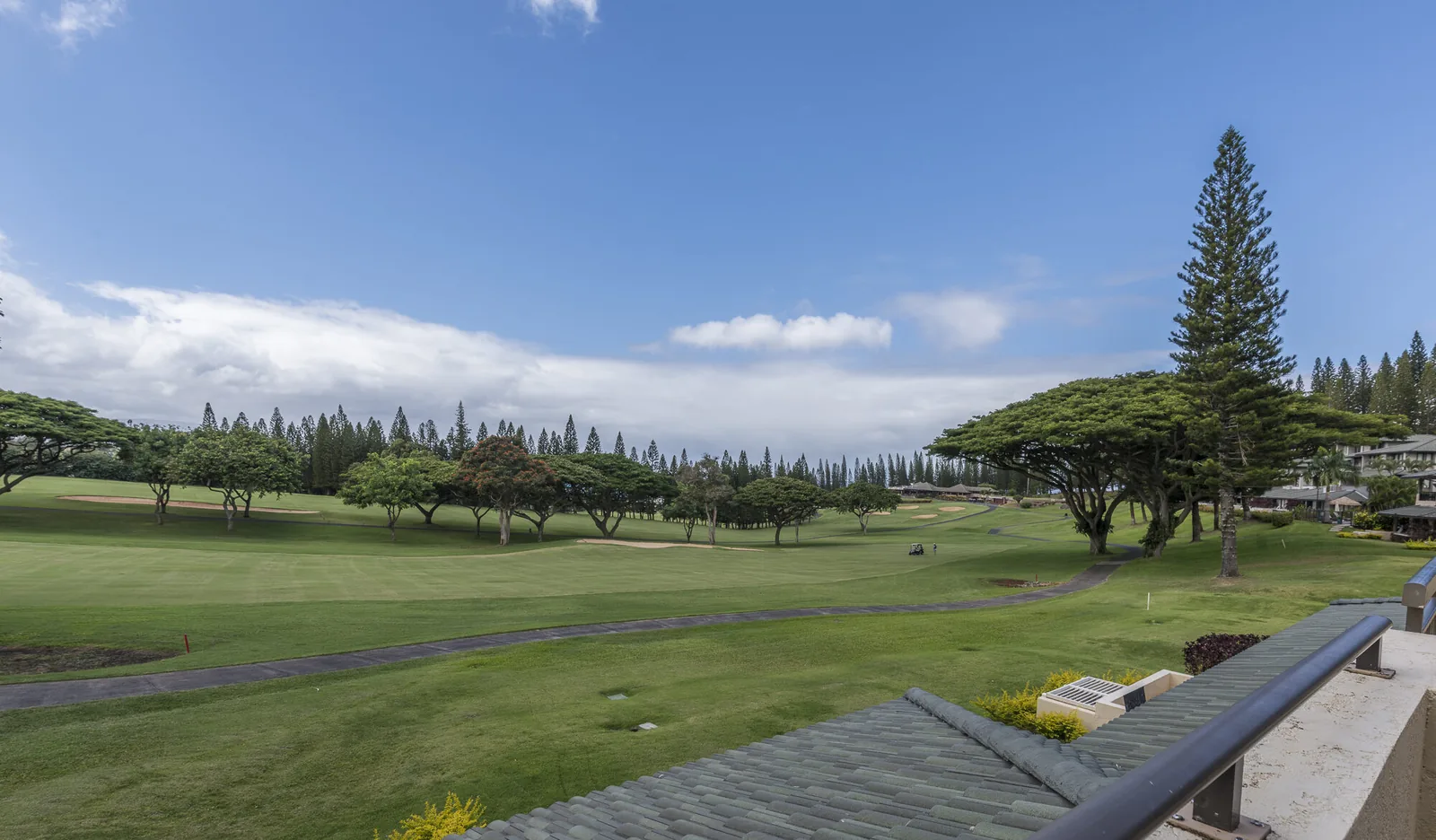 View from a Kapalua Golf Villa lanai over a golf fairway with bunkers, a cart path, Cook pines and a single golf cart.