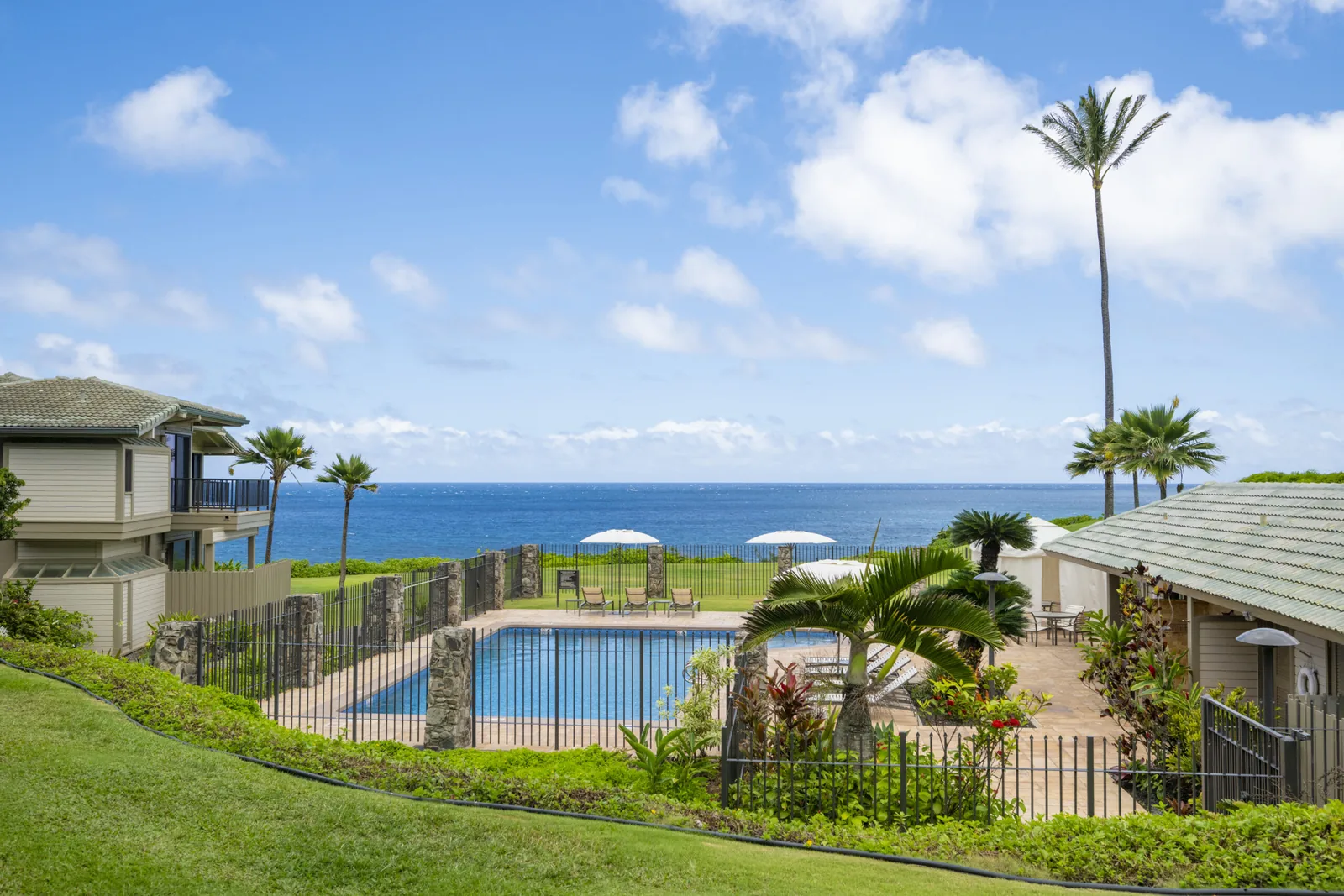 Kapalua Bay Villas grounds: low plantation-style buildings with green tile roofs, a fenced pool with white umbrellas, lawn, palms and an open ocean horizon.